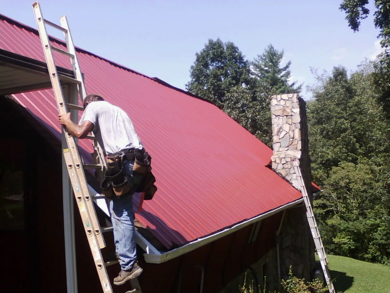 Roofer on a ladder beside a red metal roof and stone chimney — Soffit & Fascia Repair by Roofing Brothers in Oregonia